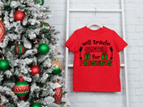 Red t-shirt with Christmas-themed text next to a decorated Christmas tree on a white brick wall.