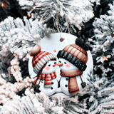 Decorative ornament with two snowpeople wearing hats and scarves on a snowy tree background