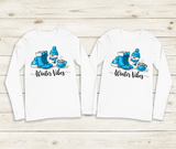 Two white long-sleeve shirts with winter-themed designs on a wooden surface.