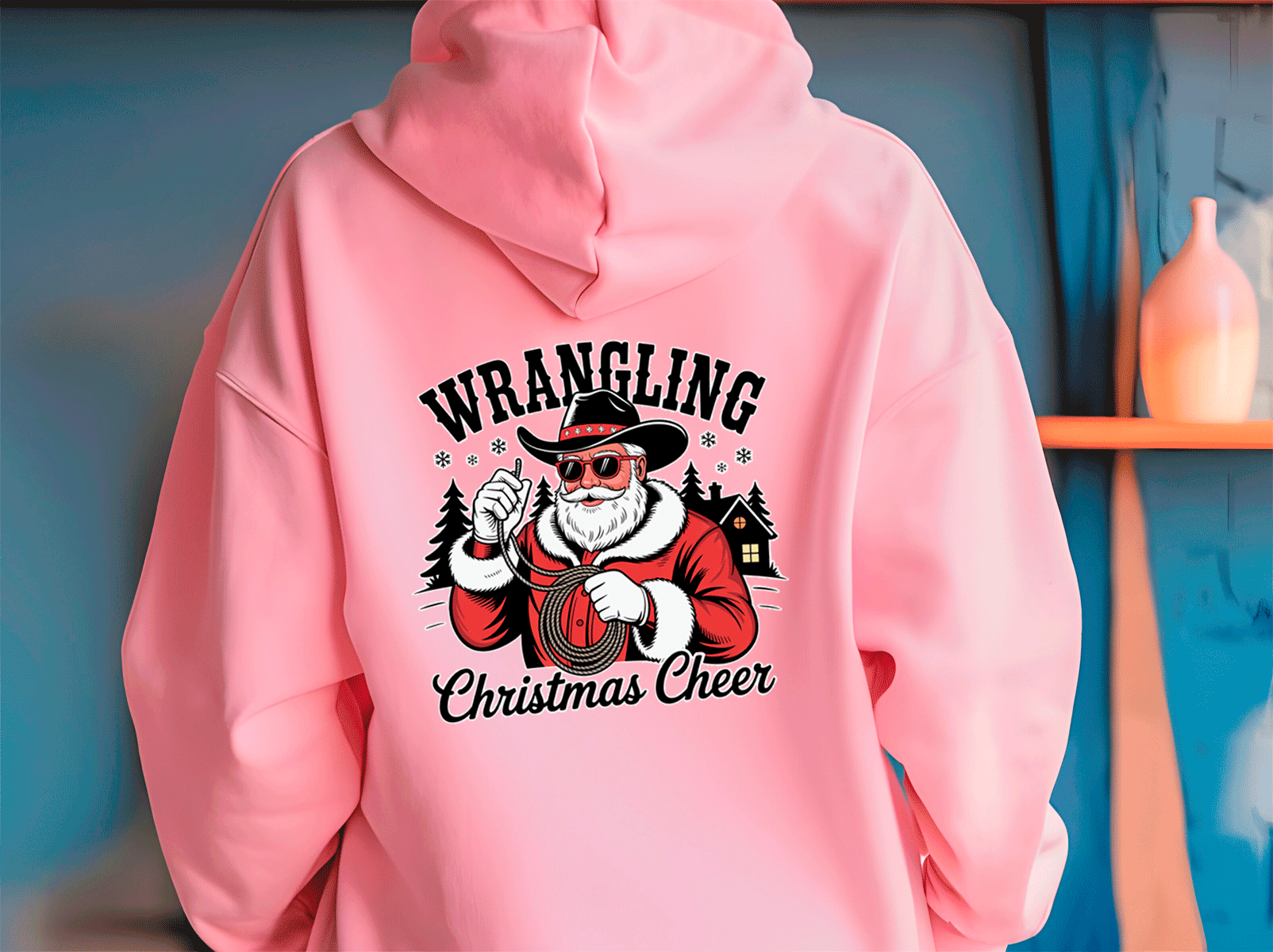 Pink hoodie with 'Wrangling Christmas Cheer' design on a blue wall background