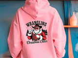 Pink hoodie with 'Wrangling Christmas Cheer' design on a blue wall background